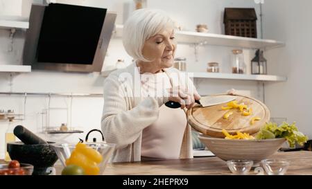 femme âgée positive préparant une salade de légumes et ajoutant du poivron en tranches dans un bol Banque D'Images