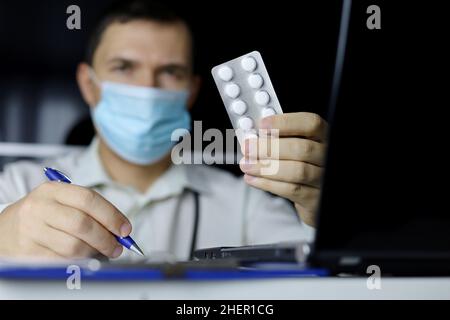 Pilules en main du médecin dans le masque médical assis à l'ordinateur portable dans le bureau.Le médecin écrit la prescription, le concept de médicaments, antibiotique ou vitamine Banque D'Images