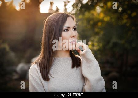 Femme pensive avec la main près du visage dans le parc à l'extérieur parmi les arbres au coucher du soleil.Photo de haute qualité Banque D'Images