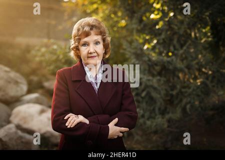 Belle femme caucasienne adulte avec des cheveu blond courts et des bras croisés dans le parc avec un manteau bordeaux et une écharpe.Photo de haute qualité Banque D'Images