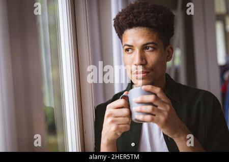 Jeune homme biracial attentionné avec tasse à café tout en regardant par la fenêtre à la maison Banque D'Images