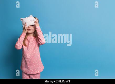 une fille en pyjama sur fond bleu sourit et ferme un œil avec un masque de sommeil Banque D'Images