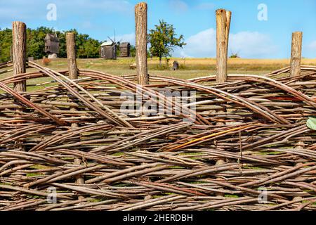 Clôture rurale composée de branches minces entrelacées sur fond de champ agricole et de moulins à vent en flou. Banque D'Images