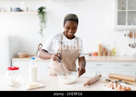 Joyeux jeune afro-américaine en tablier faire la pâte pour la pâtisserie, prépare manger dans la cuisine scandinave Banque D'Images