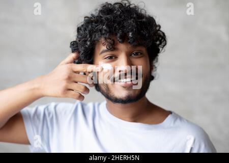 Beau homme indien appliquant de la crème hydratante sur le visage tout en se tenant dans la salle de bains, faisant la routine du matin Banque D'Images