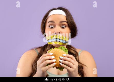 Femme européenne millénaire choquée plus taille en uniforme de sport avec col fermé avec mètre ruban regarder le hamburger Banque D'Images