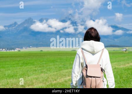 Une femme touriste dans un sweat à capuche blanc et un sac à dos bénéficie d'une belle vue sur les montagnes de Tatra tout en se tenant dans un champ vert.Les nuages flottent sur les sommets de montagne, l'espace de copie Banque D'Images