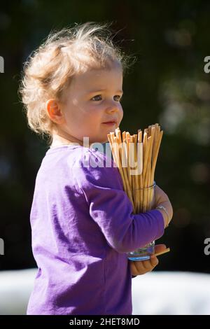 le joli bébé fille aux cheveux blonds tient le verre avec des bâtons de sel de bretzel Banque D'Images