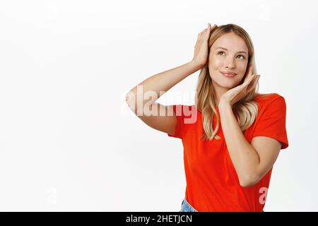 Soins de la peau et beauté des femmes.Belle fille moderne aux cheveux blonds, tenant les mains près de son visage et de la tête, souriant heureux, regardant en haut à gauche Banque D'Images