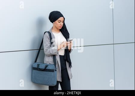 Vue de face d'une femme musulmane portant un foulard et tenant son sac à main debout en utilisant son téléphone contre un mur blanc avec une ligne noire Banque D'Images