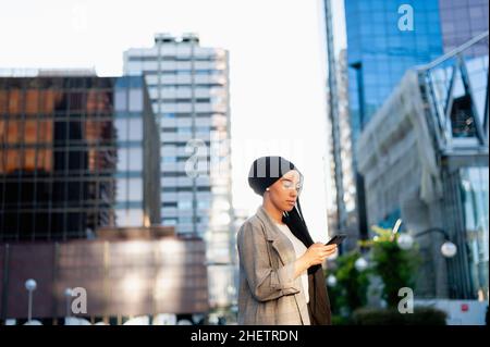 Vue latérale d'une femme musulmane souriante avec un foulard noir debout à l'aide de son téléphone dans une zone urbaine par temps ensoleillé. Banque D'Images