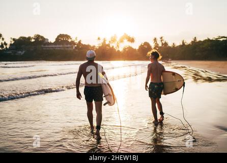 Père avec un fils adolescent marchant avec des planches de surf près de la plage de sable de l'océan avec des palmiers sur fond éclairé par le soleil de coucher de soleil.Ils sourient et ha Banque D'Images