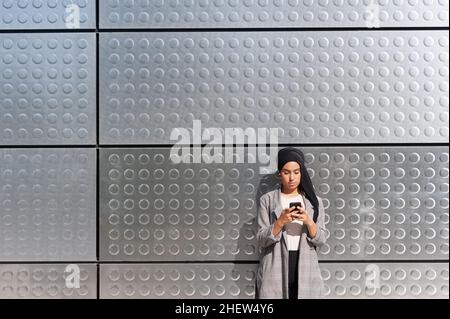 Vue latérale d'une femme musulmane debout avec son téléphone et un mur de panneaux de fer avec des motifs circulaires. Banque D'Images