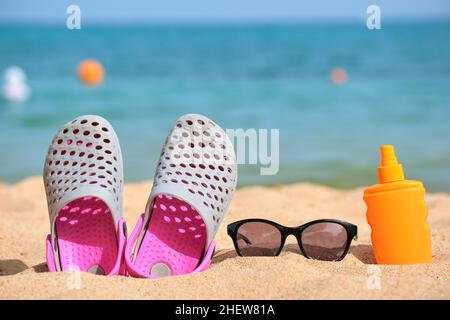 Gros plan sur des chaussures de sabots, de la crème solaire et des lunettes de soleil noires de protection sur une plage de sable au bord de la mer tropicale par temps chaud et ensoleillé. Concept de vacances d'été. Banque D'Images