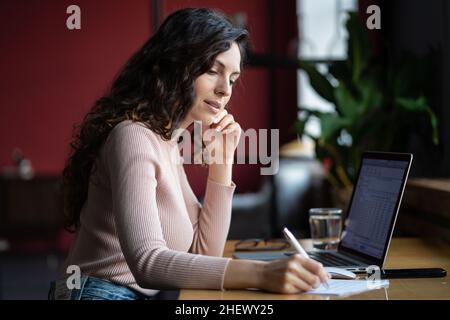 Femme d'affaires concentrée remplissant un document ou un formulaire fiscal, tout en travaillant sur un ordinateur portable au bureau Banque D'Images