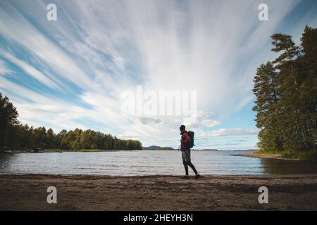 Le randonneur portant une veste et portant un sac à dos marche le long de la plage et regarde le lac Jatkonjarvi au coucher du soleil dans le parc national de Koli, dans l'est de la Finlande.Ma Banque D'Images