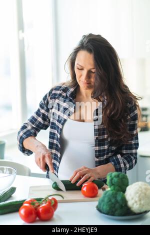 Une femme enceinte méconnaissable prépare une salade de concombre sur une planche à découper en bois avec des légumes frais et des fruits sur une table dans la cuisine.Grossesse, compagnon Banque D'Images