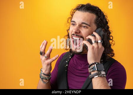 Un jeune homme aux cheveux longs et bouclés parle émotionnellement sur un téléphone portable sur fond jaune Banque D'Images