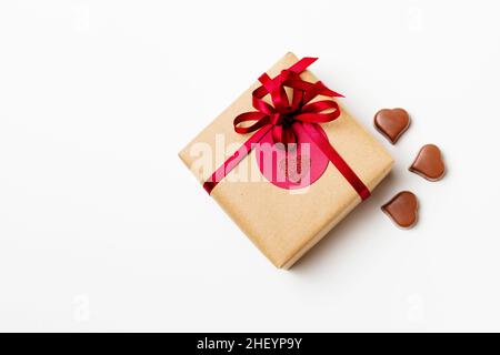 Boîte cadeau romantique enveloppée de papier artisanal et d'un noeud rouge avec bonbons au chocolat en forme de cœur sur fond blanc Banque D'Images