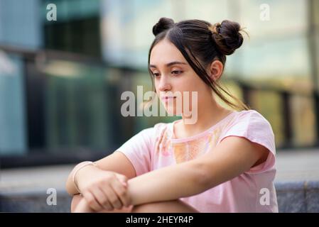 Une jeune fille adolescente pensive avec des petits pains à cheveux assis seul dans la rue et pensant à la vie sur fond flou Banque D'Images
