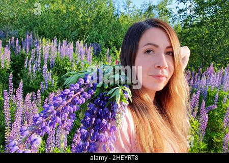 Portrait en gros plan de la jeune femme caucasienne avec de longs cheveux rouges, sur fond de verdure dans le champ de lupin.Belle cueillette de fleurs fémorales Banque D'Images