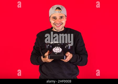 Homme dans une casquette vêtue de noir, tient un téléphone rétro du 60s et sourit, sur un fond rouge, avec un espace de copie.Concept de contact, relat social Banque D'Images