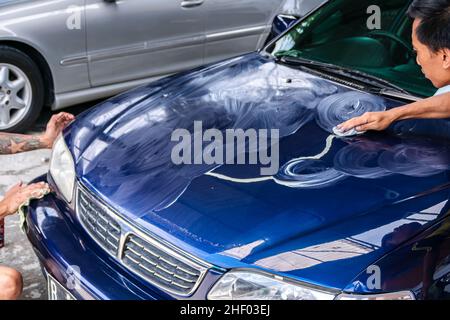 deux hommes frottent et polissent le capot de voiture bleu ou le capot moteur avec la main pour obtenir propre , brillant et brillant au garage pour les travaux de peinture et de détail automobile Banque D'Images