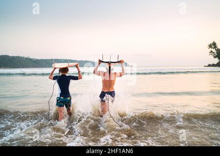 Jeune garçon avec son père avec des planches de surf sur la tête marchant dans les vagues sur la plage d'Udawalawe au Sri Lanka.Vacances actives en famille ou sport actif pe Banque D'Images