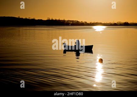 Une silhouette de pêcheur dans un petit bateau sur un réservoir au coucher du soleil avec le soleil couchant se reflétant sur l'eau. Banque D'Images