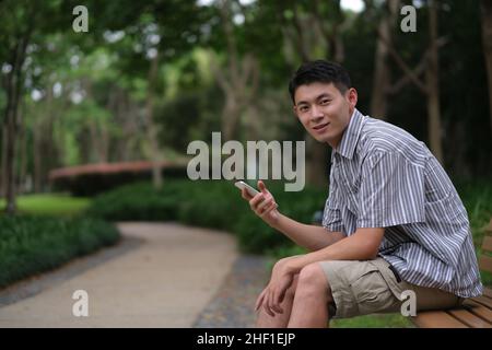 Un jeune homme asiatique souriant et utilisant le téléphone, regardant la caméra dans le parc naturel vert Banque D'Images