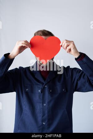 Portrait d'un homme barbu, d'un réparateur, d'un ingénieur ou d'un mécanicien en uniforme bleu debout près du mur tenant la face couvrant en forme de cœur rouge.Concept de la Saint-Valentin.Image de haute qualité Banque D'Images