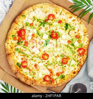 Grande pizza italienne naturelle au pesto avec pâte fine, poulet, mozzarella, tomates cerises et bazil dans une boîte artisanale avec feuilles de palmier.Cuisine méditerranéenne Banque D'Images