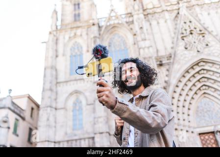 De dessous de la bonne barbe homme blogueur enregistrement vidéo sur téléphone portable avec microphone tout en se tenant près de la cathédrale historique pendant le voyage Banque D'Images