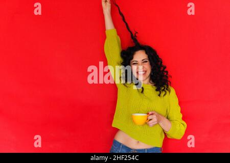 Ambiance ludique.Portrait studio de jeune femme joyeuse avec tasse de thé dans les mains jouant avec ses cheveux bouclés et souriant gaiement à l'appareil photo, isola Banque D'Images