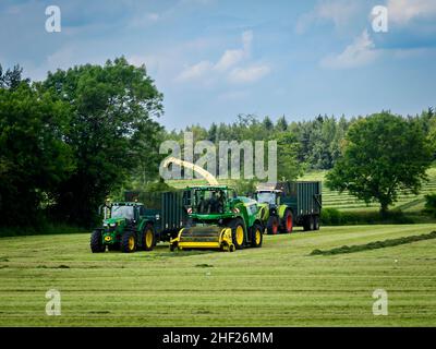 2 deux tracteurs haymaking, travaillant dans les champs agricoles avec une récolteuse John Deere chargeant des remorques (ensilage d'herbe coupée) - Yorkshire, Angleterre, Royaume-Uni. Banque D'Images