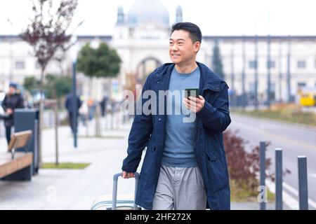 Touriste asiatique, passager de sexe masculin arrive de l'aéroport, se balade dans la ville avec une valise, un homme sourit et se réjouit utilise le téléphone et l'apple Banque D'Images
