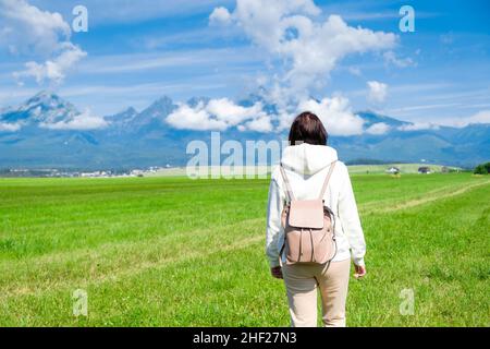 Une femme touriste dans un sweat à capuche blanc et un sac à dos bénéficie d'une belle vue sur les montagnes de Tatra tout en se tenant dans un champ vert.Les nuages flottent sur les sommets de montagne, l'espace de copie Banque D'Images