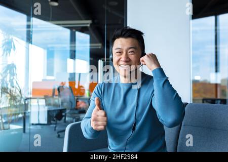 Souriant et réjouissant, homme d'affaires travaillant pour des appels vidéo, des conférences à distance, regardant la caméra et pointant les pouces vers le haut, à l'aide d'un micro-casque, Banque D'Images