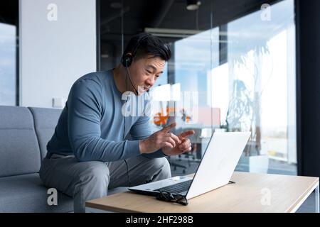 Dans un bureau moderne, un homme asiatique souriant et heureux utilise un micro-casque pour un appel vidéo, un homme d'affaires sourit à l'interlocuteur et regarde la scrore de l'ordinateur portable Banque D'Images