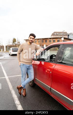 Jeune latino ou arabe est homme debout près de petite voiture rouge sur la fente de stationnement Banque D'Images