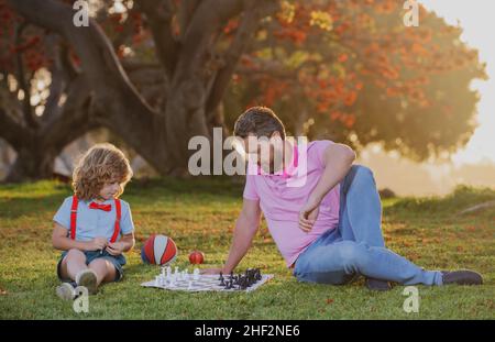 École d'échecs pour enfants.Père et fils jouant aux échecs allongé sur l'herbe au parc de pelouse.Fête des pères, famille d'amour, parentalité, concept d'enfance.Enfant concentré Banque D'Images