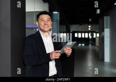 Un homme d'affaires asiatique prospère, le patron, dans un costume d'affaires se réjouit et sourit utilise une tablette, travaille dans un bureau moderne Banque D'Images