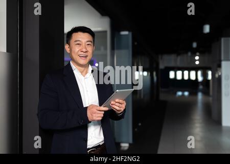 Un homme d'affaires asiatique prospère, le patron, dans un costume d'affaires se réjouit et sourit utilise une tablette, travaille dans un bureau moderne Banque D'Images