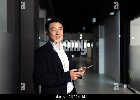 Un homme d'affaires asiatique prospère, le patron, dans un costume d'affaires se réjouit et sourit utilise une tablette, travaille dans un bureau moderne Banque D'Images