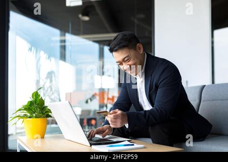 Le patron asiatique transfère de l'argent d'une carte bancaire, assis dans le bureau, satisfait du résultat souriant d'homme d'affaires, travaille sur ordinateur portable Banque D'Images