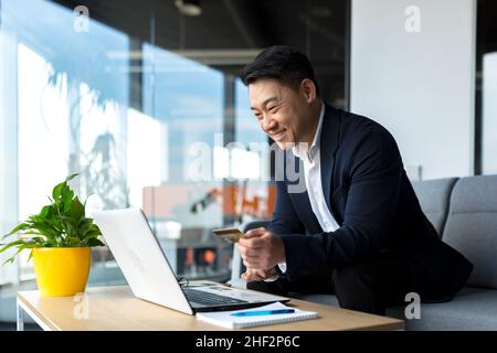 Le patron asiatique transfère de l'argent d'une carte bancaire, assis dans le bureau, satisfait du résultat souriant d'homme d'affaires, travaille sur ordinateur portable Banque D'Images