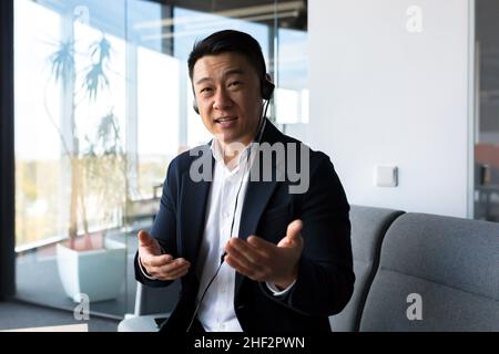 Asiatique amical regarder la webcam et parler, homme d'affaires avec le casque souriant à l'interlocuteur, appel vidéo de bureau moderne Banque D'Images