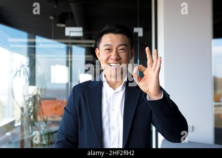 Homme d'affaires asiatique heureux regardant l'appareil photo montrant les pouces vers le haut et le signe ok, homme d'affaires souriant dans le bureau moderne Banque D'Images