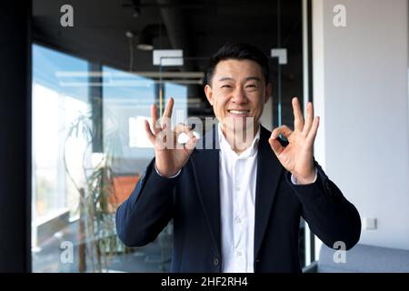 Homme d'affaires asiatique heureux regardant l'appareil photo montrant les pouces vers le haut et le signe ok, homme d'affaires souriant dans le bureau moderne Banque D'Images