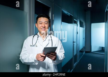 Portrait d'un médecin asiatique joyeux et joyeux, homme dans une clinique moderne regardant l'appareil photo et souriant tenant un ordinateur tablette Banque D'Images